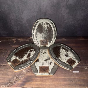 May include: A set of four horseshoe-shaped coasters with a cowhide pattern. Each coaster has a brown leather patch with a logo. The coasters are arranged on a wooden surface. The text "Set # 101" is visible.