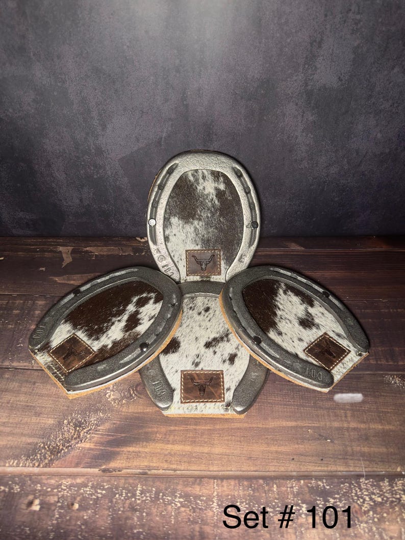 May include: A set of four horseshoe-shaped coasters with a cowhide pattern. Each coaster has a brown leather patch with a logo. The coasters are arranged on a wooden surface. The text "Set # 101" is visible.