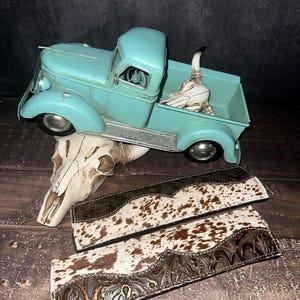 May include: A turquoise vintage pickup truck with a decorative skull in the truck bed. Two leather belts with a cowhide pattern and embossed floral designs are displayed on a wooden surface. A decorative skull is also present.