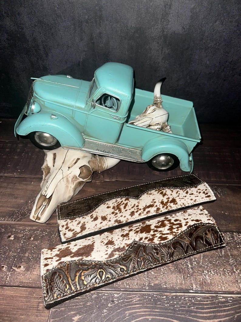 May include: A turquoise vintage pickup truck with a decorative skull in the truck bed. Two leather belts with a cowhide pattern and embossed floral designs are displayed on a wooden surface. A decorative skull is also present.