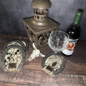 May include: A rustic still life featuring horseshoe-shaped coasters with cowhide centers, a decorative lantern, a wine glass, a bottle of wine, and a small skull. The coasters have a leather patch with a logo.