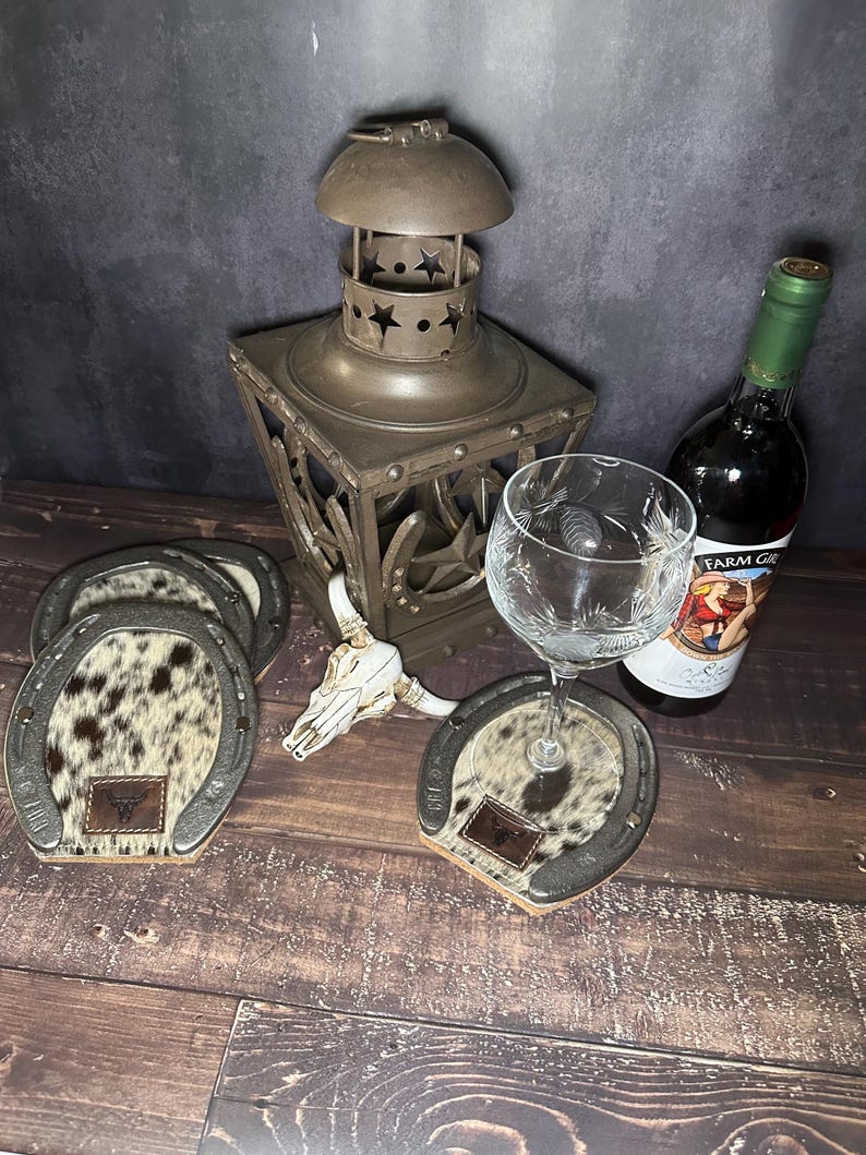 May include: A rustic still life featuring horseshoe-shaped coasters with cowhide centers, a decorative lantern, a wine glass, a bottle of wine, and a small skull. The coasters have a leather patch with a logo.