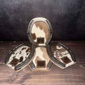 May include: Set of four horseshoe-shaped coasters. Each coaster features a silver-colored metal frame and a cowhide center with brown and white patterns. A small, brown leather patch with a logo is centered on each coaster. The text "Set# H105" is at the bottom.
