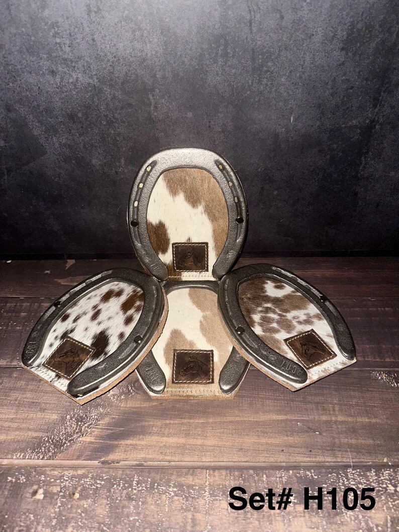 May include: Set of four horseshoe-shaped coasters. Each coaster features a silver-colored metal frame and a cowhide center with brown and white patterns. A small, brown leather patch with a logo is centered on each coaster. The text "Set# H105" is at the bottom.