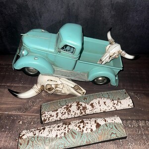 May include: A turquoise metal vintage truck model with a white cow skull in the bed. Two additional cow skulls and two leather bands with a turquoise and brown cowhide pattern are also displayed.