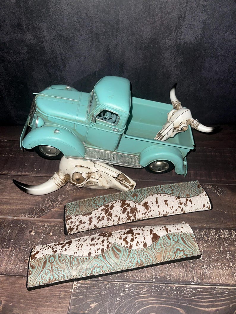 May include: A turquoise metal vintage truck model with a white cow skull in the bed. Two additional cow skulls and two leather bands with a turquoise and brown cowhide pattern are also displayed.