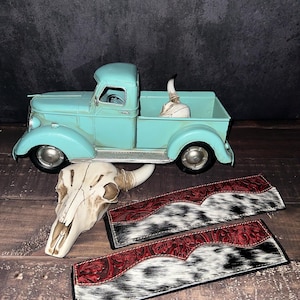 May include: A turquoise vintage pickup truck model with a cow skull in the bed. Two decorative leather pieces with red tooled leather and black and white cowhide are in the foreground. A second cow skull is also present.