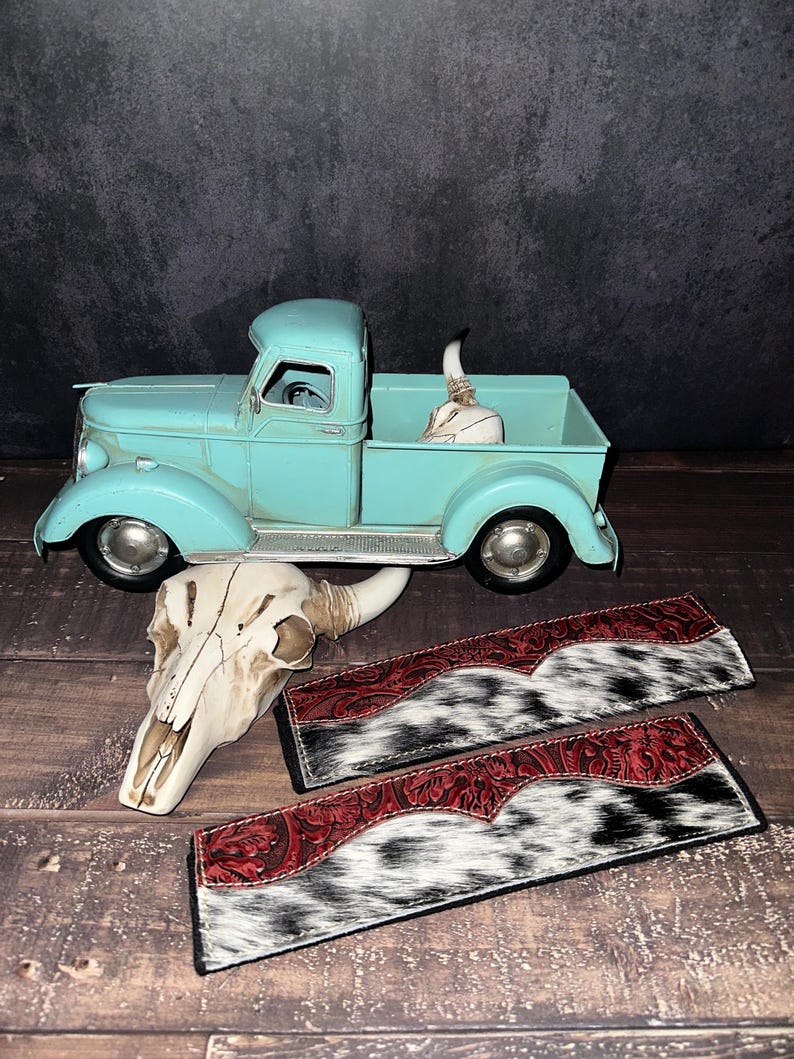 May include: A turquoise vintage pickup truck model with a cow skull in the bed. Two decorative leather pieces with red tooled leather and black and white cowhide are in the foreground. A second cow skull is also present.