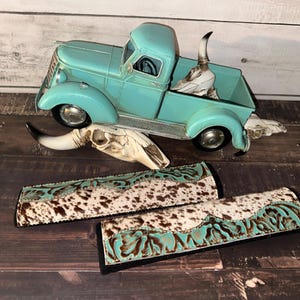 May include: A turquoise vintage pickup truck model with a steer skull in the bed. Two rectangular accessories with a cowhide pattern and turquoise accents are in the foreground. The items are on a wooden surface.