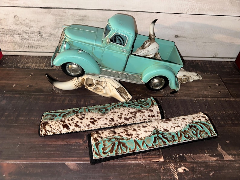 May include: A turquoise vintage pickup truck model with a steer skull in the bed. Two rectangular accessories with a cowhide pattern and turquoise accents are in the foreground. The items are on a wooden surface.