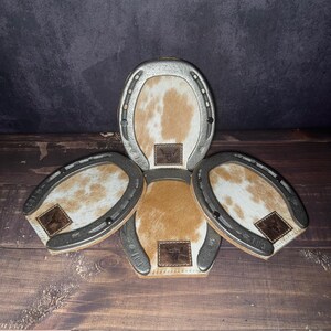 May include: A set of four horseshoe-shaped coasters. The coasters have a silver metal frame and a cowhide center with brown and white patterns. Each coaster has a small leather patch with a bull logo. The text "Set # 102" is at the bottom.
