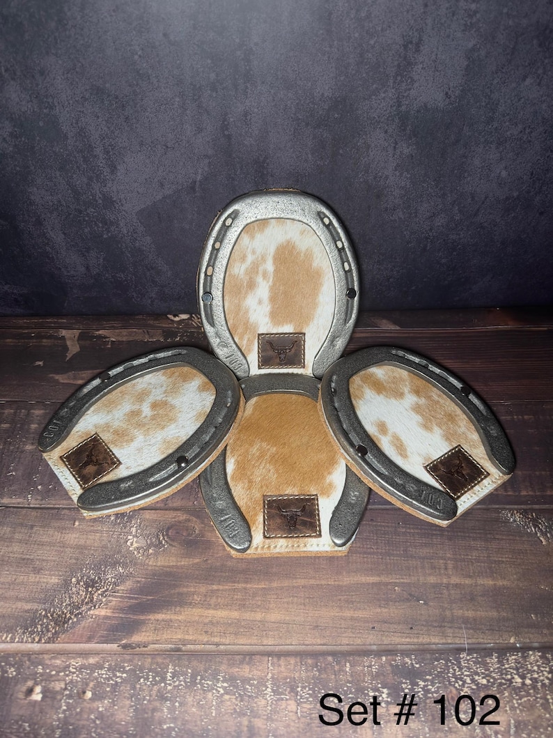 May include: A set of four horseshoe-shaped coasters. The coasters have a silver metal frame and a cowhide center with brown and white patterns. Each coaster has a small leather patch with a bull logo. The text "Set # 102" is at the bottom.