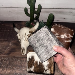 May include: Three cowhide wallets in shades of brown, white, and gray. The wallets are rectangular with stitched edges. A hand holds one wallet, with a cactus and a cow skull in the background.