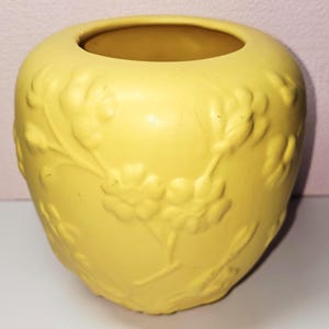 May include: A round, matte yellow ceramic vase featuring a raised, embossed floral pattern across its exterior surface. The vase has a wide, circular opening at the top and a smooth, consistent yellow finish throughout its rounded, bulbous body.