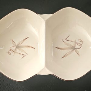 May include: A cream-colored ceramic divided serving dish with a symmetrical, elongated diamond shape. Each of the two sections features a brown, hand-painted botanical illustration of a leaf with a curled vine stem centered on the interior base.