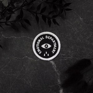 Puede incluir: Parche bordado blanco con un ojo negro y tres lágrimas negras. El texto "Emotional Department" está escrito en un círculo alrededor del ojo.