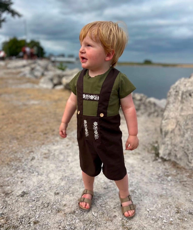 Puede incluir: Una camisa de manga corta verde oliva usada debajo de un peto marr&oacute;n oscuro con tirantes. El peto presenta un bordado floral blanco en el pecho y dos peque&ntilde;os botones de color claro en la cintura. El conjunto se combina con sandalias de color beige.