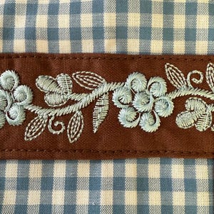Puede incluir: Un art&iacute;culo textil con un patr&oacute;n de cuadros vichy en azul claro y blanco, dividido por una banda de tela marr&oacute;n horizontal. La banda marr&oacute;n est&aacute; decorada con bordados florales en azul claro, mostrando p&eacute;talos y hojas cosidos en detalle.