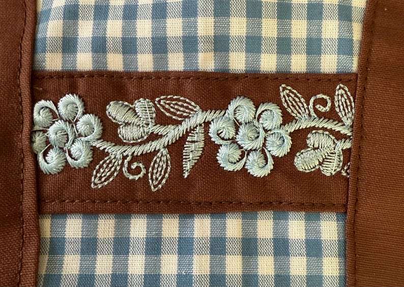 Puede incluir: Un art&iacute;culo textil con un patr&oacute;n de cuadros vichy en azul claro y blanco, dividido por una banda de tela marr&oacute;n horizontal. La banda marr&oacute;n est&aacute; decorada con bordados florales en azul claro, mostrando p&eacute;talos y hojas cosidos en detalle.