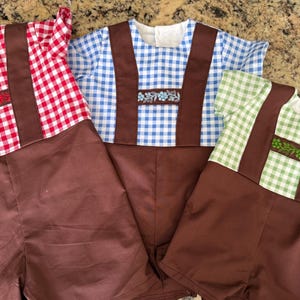 Puede incluir: Tres peleles de manga corta con pantalones cortos marrones y tirantes marrones sobre tops de cuadros vichy. Las camisas de cuadros rojos, azules y verdes tienen una banda horizontal en el pecho con bordados florales a juego en cada color.