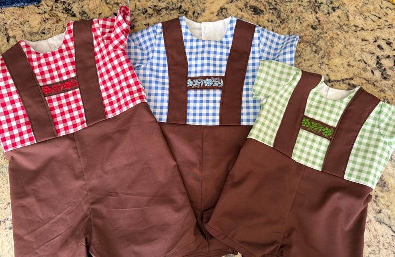 Puede incluir: Tres peleles de manga corta con pantalones cortos marrones y tirantes marrones sobre tops de cuadros vichy. Las camisas de cuadros rojos, azules y verdes tienen una banda horizontal en el pecho con bordados florales a juego en cada color.