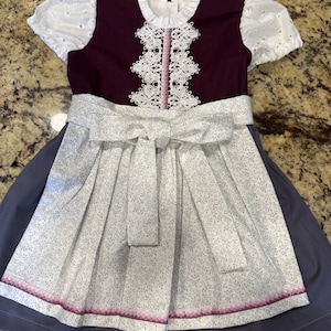Pode incluir: Um vestido de estilo tradicional com um corpete roxo escuro e uma gola de renda branca. O vestido apresenta um avental branco com um padr&atilde;o floral e um la&ccedil;o a condizer. A saia &eacute; de cor cinza-azul s&oacute;lida, e o vestido tem mangas curtas brancas com ilh&oacute;s.