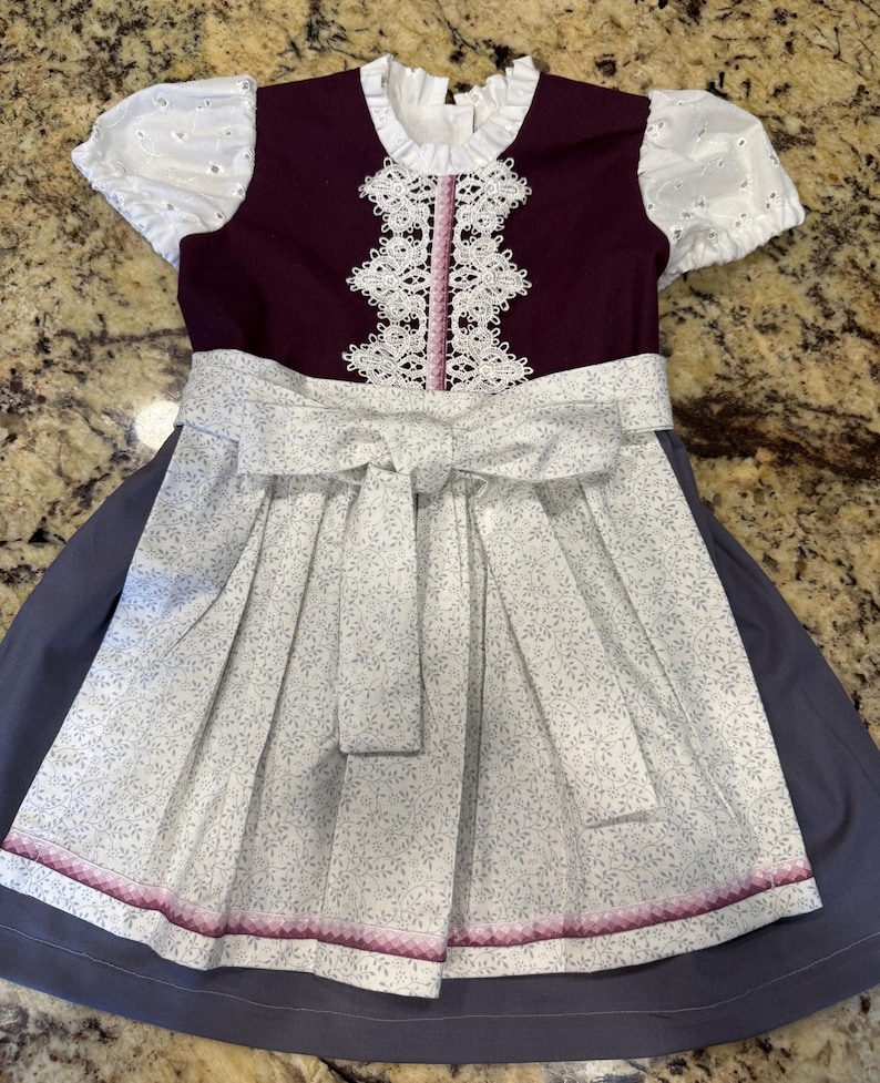 Pode incluir: Um vestido de estilo tradicional com um corpete roxo escuro e uma gola de renda branca. O vestido apresenta um avental branco com um padr&atilde;o floral e um la&ccedil;o a condizer. A saia &eacute; de cor cinza-azul s&oacute;lida, e o vestido tem mangas curtas brancas com ilh&oacute;s.