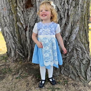 Pode incluir: Uma crian&ccedil;a pequena vestindo um vestido azul e branco com uma sobreposi&ccedil;&atilde;o de renda. O vestido tem mangas curtas e um detalhe de fita na cintura. A crian&ccedil;a tamb&eacute;m est&aacute; usando meias brancas e sapatos pretos. Um la&ccedil;o branco adorna o cabelo da crian&ccedil;a.