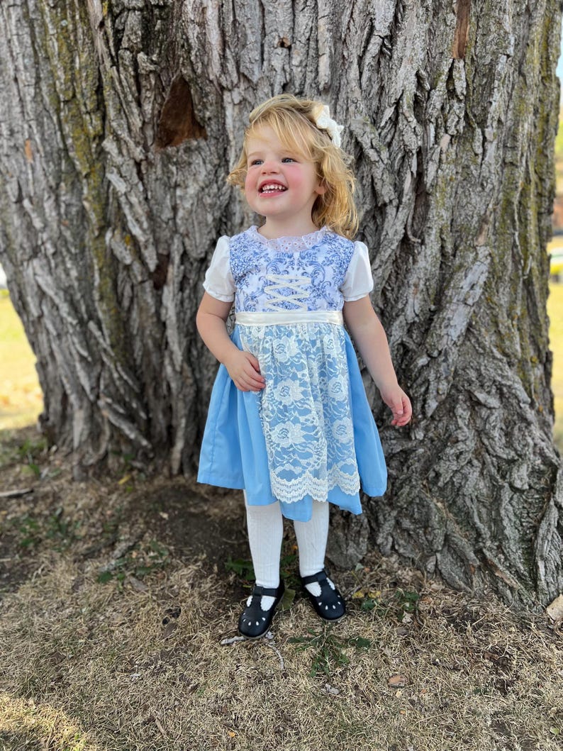 Pode incluir: Uma crian&ccedil;a pequena vestindo um vestido azul e branco com uma sobreposi&ccedil;&atilde;o de renda. O vestido tem mangas curtas e um detalhe de fita na cintura. A crian&ccedil;a tamb&eacute;m est&aacute; usando meias brancas e sapatos pretos. Um la&ccedil;o branco adorna o cabelo da crian&ccedil;a.