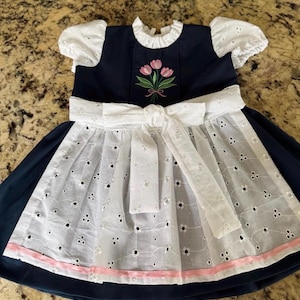 Puede incluir: Un vestido azul marino con mangas abullonadas de encaje blanco y un delantal blanco a juego atado con un lazo en la cintura. El corpi&ntilde;o muestra un ramo bordado de tres tulipanes rosas con tallos verdes, y el dobladillo del delantal tiene un borde de cinta rosa claro.