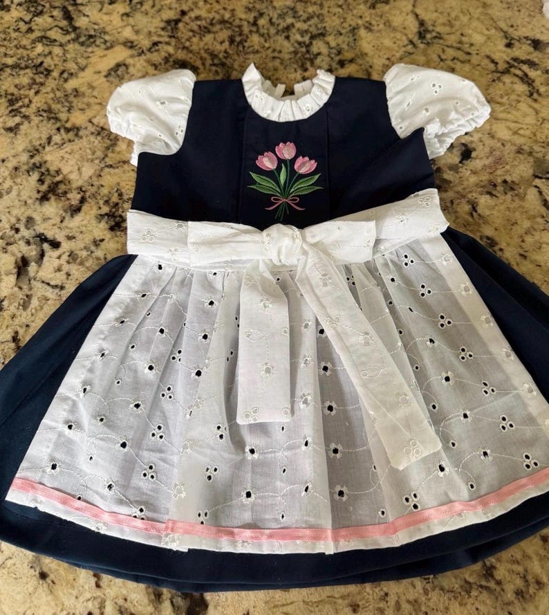 Puede incluir: Un vestido azul marino con mangas abullonadas de encaje blanco y un delantal blanco a juego atado con un lazo en la cintura. El corpi&ntilde;o muestra un ramo bordado de tres tulipanes rosas con tallos verdes, y el dobladillo del delantal tiene un borde de cinta rosa claro.