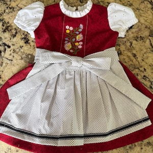 Pode incluir: Um vestido vermelho e branco com bolinhas com um grande la&ccedil;o e bordado floral. O vestido apresenta mangas brancas com ilh&oacute;s e uma gola branca. A saia tem uma faixa preta na bainha.