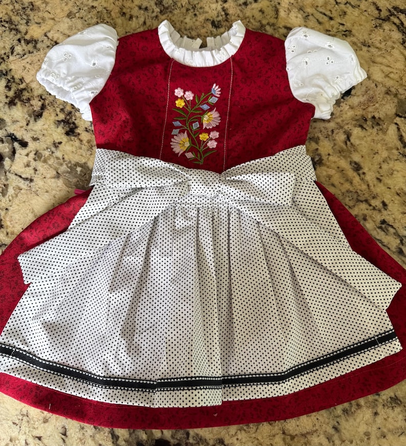 Pode incluir: Um vestido vermelho e branco com bolinhas com um grande la&ccedil;o e bordado floral. O vestido apresenta mangas brancas com ilh&oacute;s e uma gola branca. A saia tem uma faixa preta na bainha.
