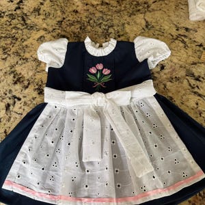 Pode incluir: Um vestido azul marinho com detalhes em ilh&oacute;s brancos e um la&ccedil;o branco. O vestido apresenta mangas curtas e bufantes brancas e um corpete bordado com tulipas rosa. Uma fita rosa adorna a bainha da saia de ilh&oacute;s.