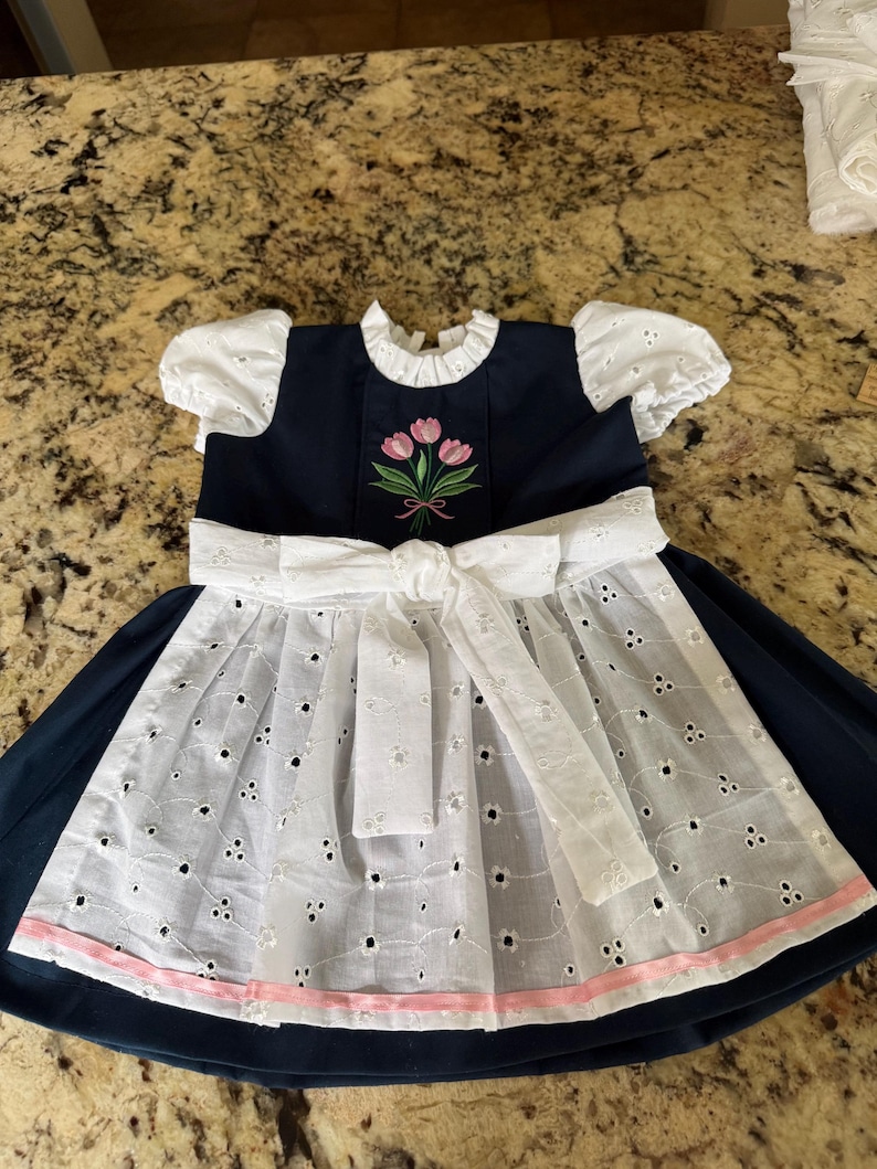 Pode incluir: Um vestido azul marinho com detalhes em ilh&oacute;s brancos e um la&ccedil;o branco. O vestido apresenta mangas curtas e bufantes brancas e um corpete bordado com tulipas rosa. Uma fita rosa adorna a bainha da saia de ilh&oacute;s.