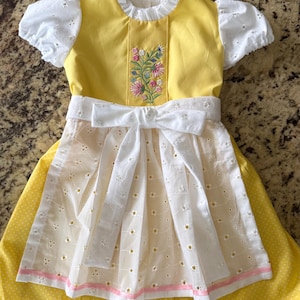 Pode incluir: Um vestido amarelo e branco com bordados florais e um la&ccedil;o branco. O vestido apresenta mangas curtas com ilh&oacute;s e um avental branco com ilh&oacute;s sobre uma saia amarela com bolinhas. O corpete &eacute; amarelo com bordados florais.