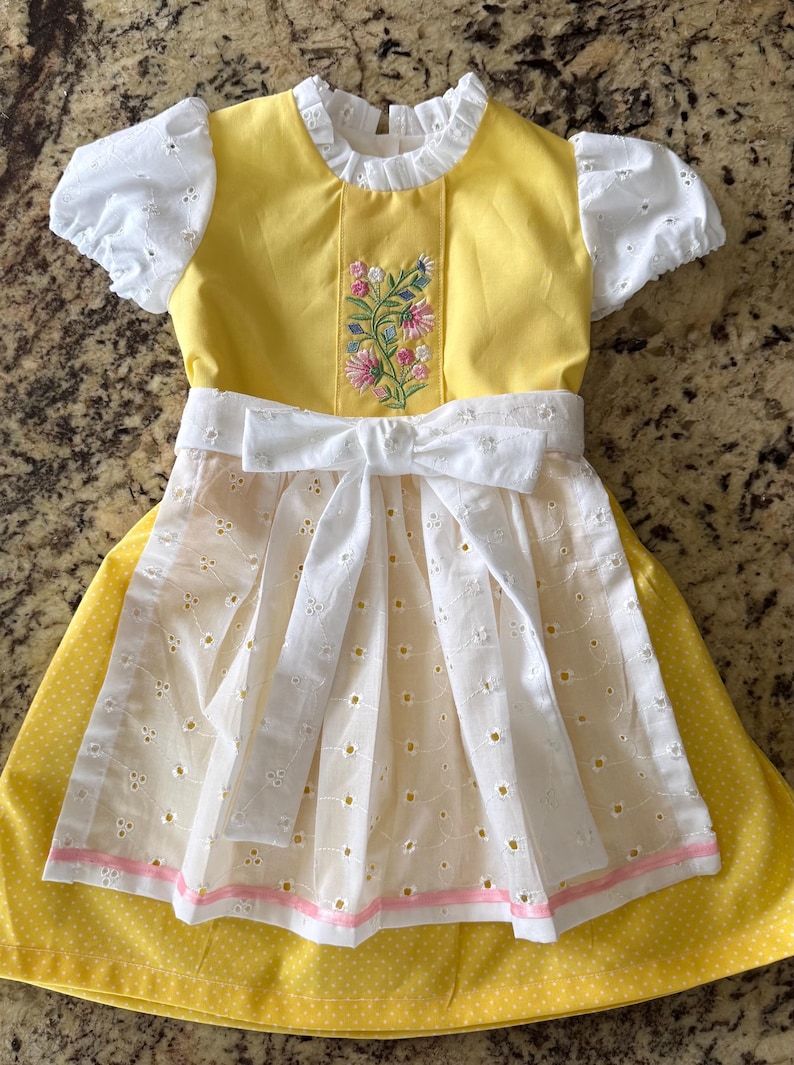 Pode incluir: Um vestido amarelo e branco com bordados florais e um la&ccedil;o branco. O vestido apresenta mangas curtas com ilh&oacute;s e um avental branco com ilh&oacute;s sobre uma saia amarela com bolinhas. O corpete &eacute; amarelo com bordados florais.