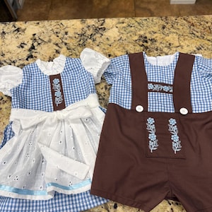 Pode incluir: Dois conjuntos a condizer: um vestido azul e branco xadrez com um avental branco com bordados e um la&ccedil;o castanho, e um conjunto de macac&atilde;o castanho com top xadrez azul e branco. Ambos os conjuntos t&ecirc;m bordados florais.