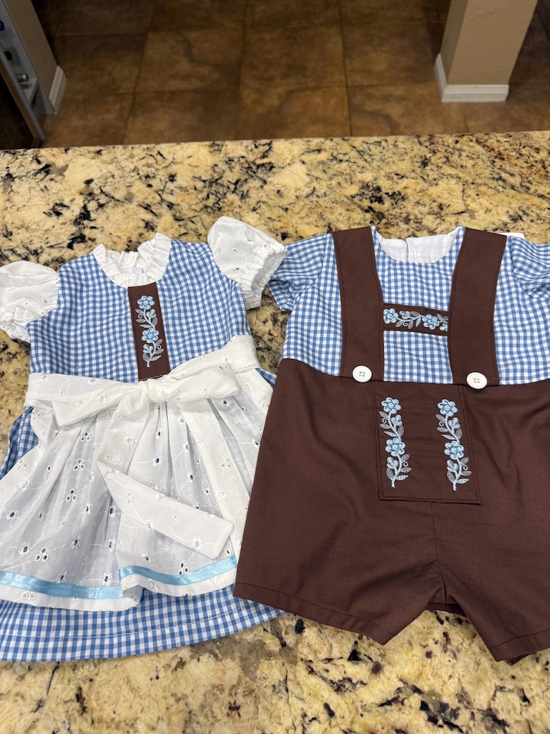 Pode incluir: Dois conjuntos a condizer: um vestido azul e branco xadrez com um avental branco com bordados e um la&ccedil;o castanho, e um conjunto de macac&atilde;o castanho com top xadrez azul e branco. Ambos os conjuntos t&ecirc;m bordados florais.