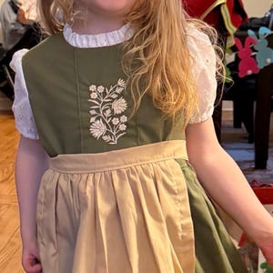 Puede incluir: Un vestido verde oliva con una blusa blanca de manga corta con bordado ingl&eacute;s debajo. El vestido tiene un delantal color canela sujeto a la cintura y un dise&ntilde;o de bordado floral blanco en el pecho. La falda tiene un borde horizontal blanco.