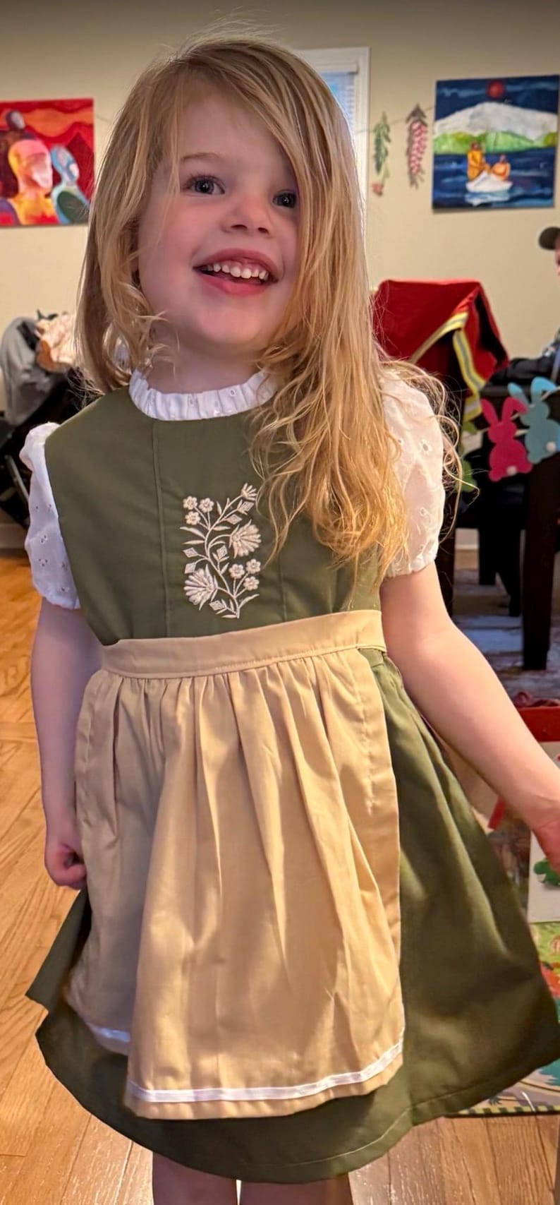 Puede incluir: Un vestido verde oliva con una blusa blanca de manga corta con bordado ingl&eacute;s debajo. El vestido tiene un delantal color canela sujeto a la cintura y un dise&ntilde;o de bordado floral blanco en el pecho. La falda tiene un borde horizontal blanco.