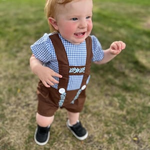May include: A toddler outfit consisting of a blue and white gingham short-sleeve shirt paired with brown suspender shorts. The brown shorts feature white buttons and light blue floral embroidery along the suspender straps and the front waistband.