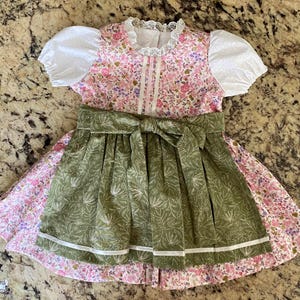 Pode incluir: Um vestido com estampa floral com um avental verde e mangas brancas. O vestido apresenta um padr&atilde;o floral rosa no corpete e na saia, com um avental verde amarrado na cintura. As mangas s&atilde;o brancas e bufantes, e o vestido tem uma gola de renda.
