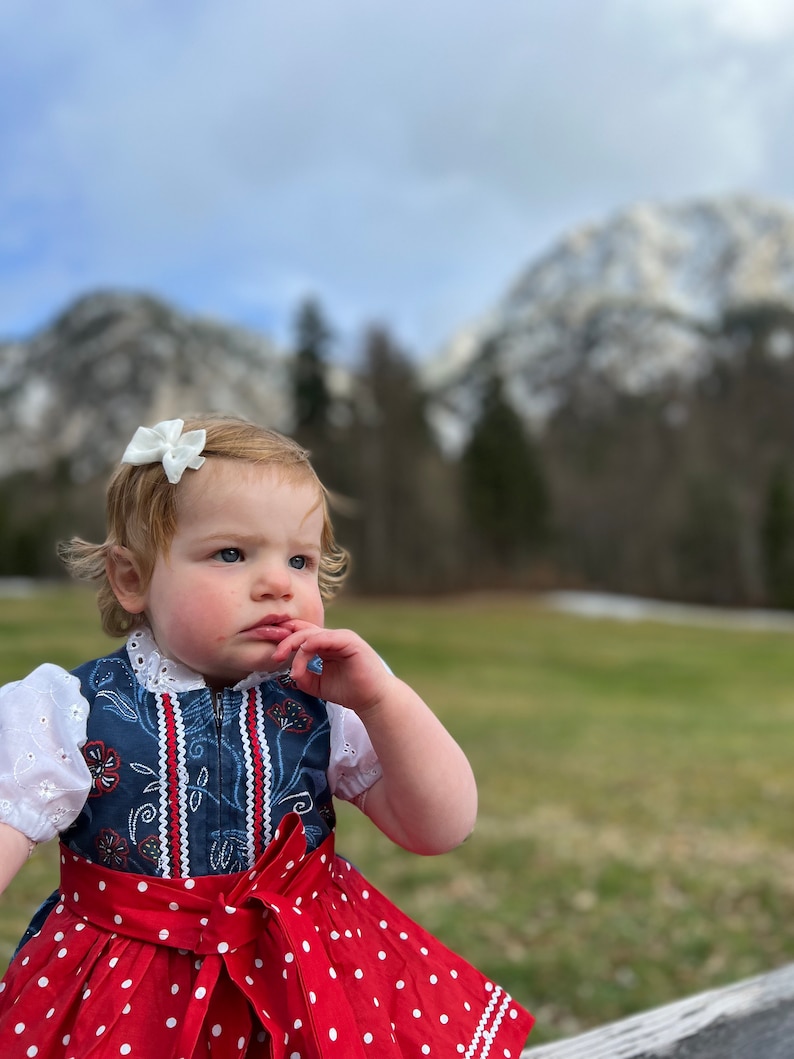 Pode incluir: Uma crian&ccedil;a pequena vestindo um tradicional vestido dirndl alem&atilde;o. O vestido apresenta um corpete azul marinho com bordados florais, mangas de ilh&oacute;s brancas e uma saia vermelha com bolinhas. Um la&ccedil;o vermelho adorna a cintura. A crian&ccedil;a tem um la&ccedil;o branco no cabelo.