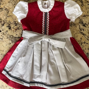 Pode incluir: Um vestido dirndl vermelho e branco com uma saia cinza de bolinhas e um la&ccedil;o grande. O vestido apresenta mangas brancas com ilh&oacute;s e uma gola branca com babados. Uma fita preta e branca adorna a bainha da saia.