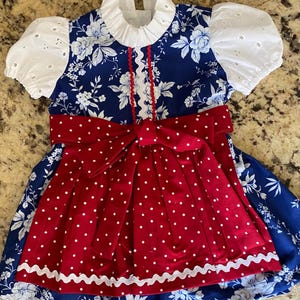 Pode incluir: Um vestido azul e branco com estampa floral com um avental e la&ccedil;o de bolinhas vermelhas. O vestido apresenta mangas de ilh&oacute;s brancas e uma gola com babados. A borda ondulada branca acentua o avental. Um vestido charmoso com tema patri&oacute;tico.