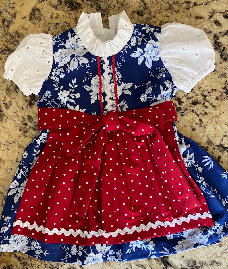 Pode incluir: Um vestido azul e branco com estampa floral com um avental e la&ccedil;o de bolinhas vermelhas. O vestido apresenta mangas de ilh&oacute;s brancas e uma gola com babados. A borda ondulada branca acentua o avental. Um vestido charmoso com tema patri&oacute;tico.