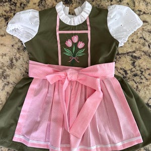Puede incluir: Un vestido verde oliva con mangas abullonadas de encaje blanco y cuello alto con volantes. El corpi&ntilde;o presenta un ramo de tulipanes rosas bordado. Un delantal rosa claro con estampado de lunares blancos y un gran lazo en la cintura.