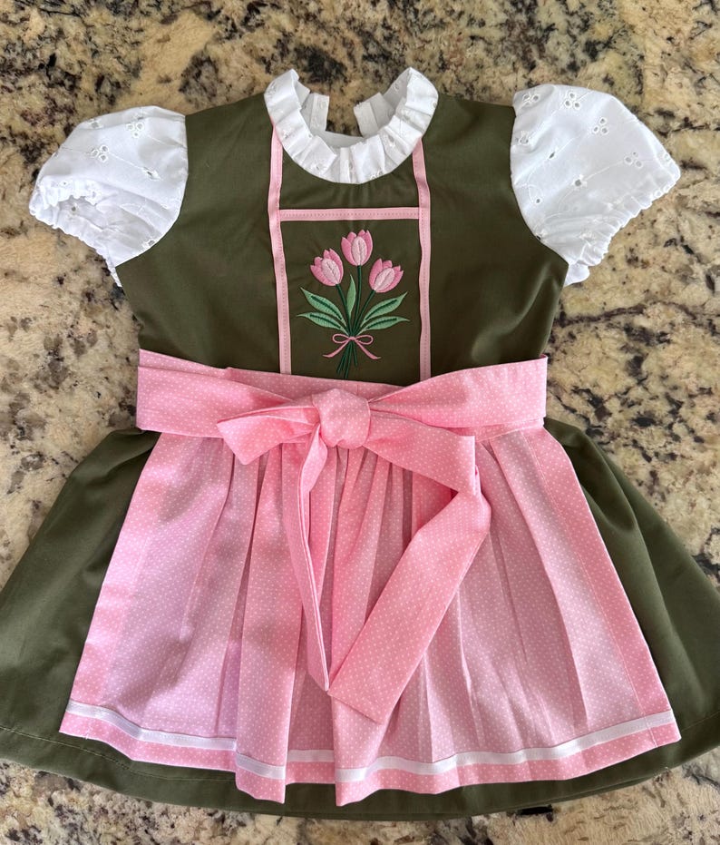 Puede incluir: Un vestido verde oliva con mangas abullonadas de encaje blanco y cuello alto con volantes. El corpi&ntilde;o presenta un ramo de tulipanes rosas bordado. Un delantal rosa claro con estampado de lunares blancos y un gran lazo en la cintura.