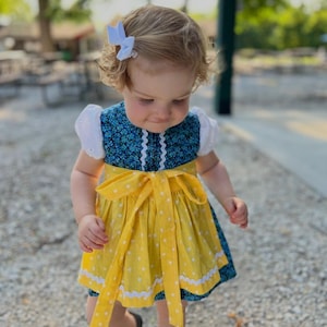 May include: A dress featuring a blue bodice with a small floral pattern, white puff sleeves, and a yellow apron overlay with white polka dots. The dress includes white rickrack trim along the bodice and the hem of the yellow apron.